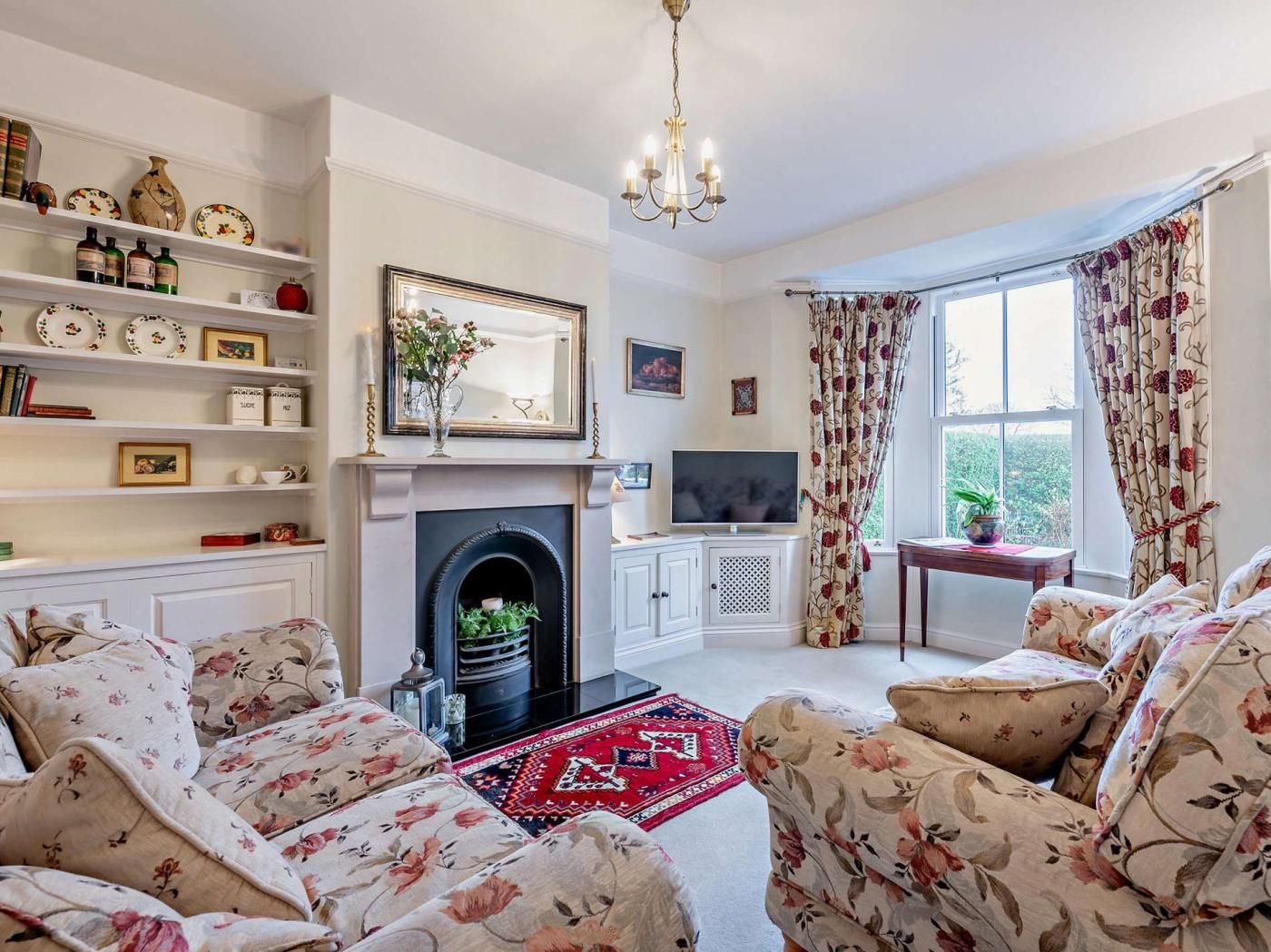 A living room with floral-patterned sofas, a fireplace, a television, and a bookshelf with decorative items.
