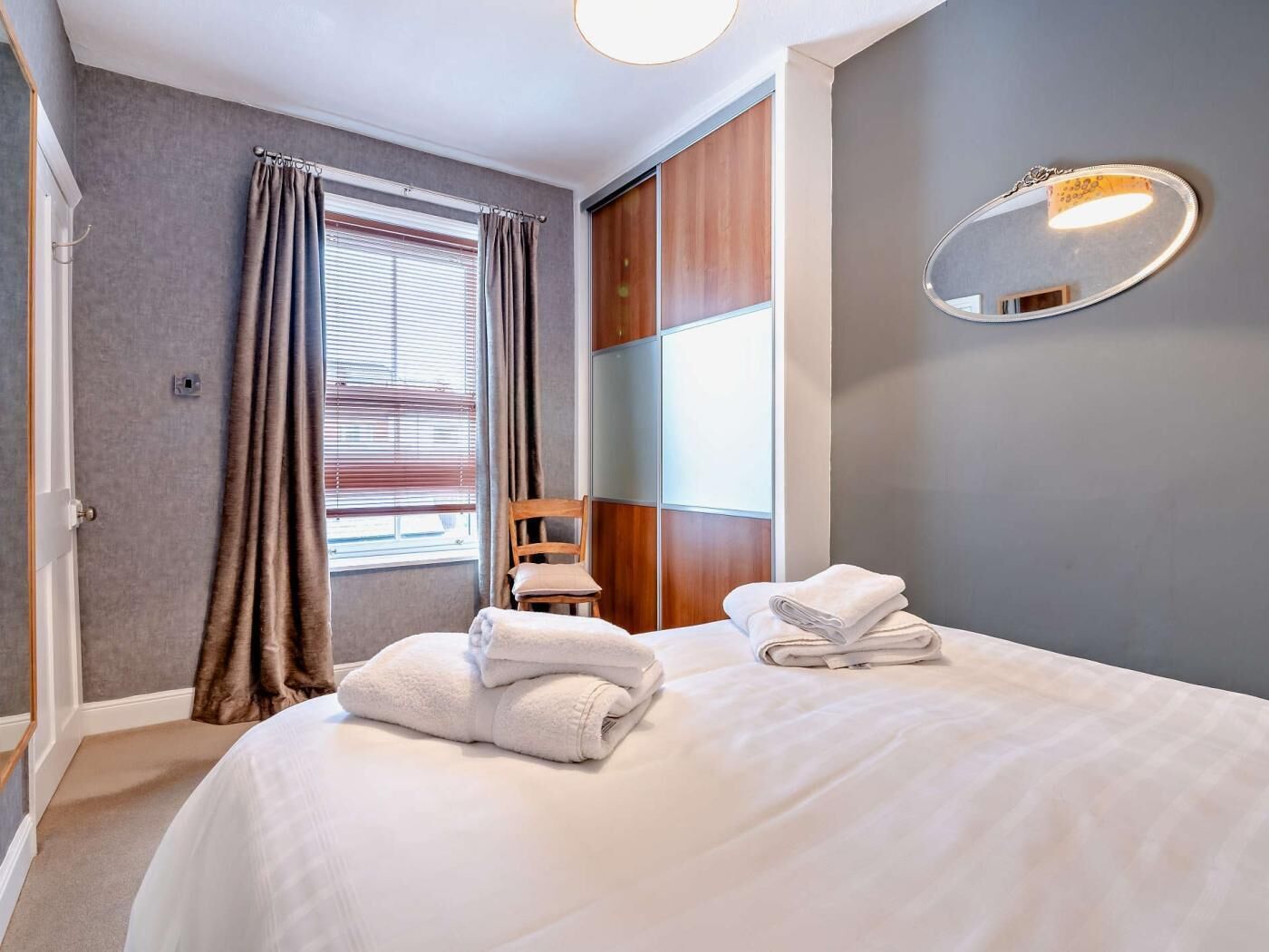 Una habitación de hotel con una cama, una silla, un espejo y una ventana con cortinas.
