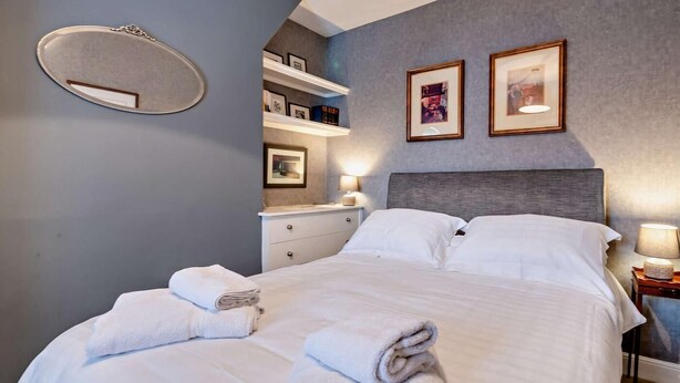 A hotel room with a bed, white towels, a bedside table with a lamp, a nightstand, and a wall-mounted mirror. - 2 Bed in Harrogate (oc-hh037) (Harrogate)