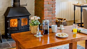 A cozy room with a wood-burning stove, a table with breakfast items, and a wicker basket with flowers. - 3 Bed in Beaworthy (oc-wvfco) (Ashwater)