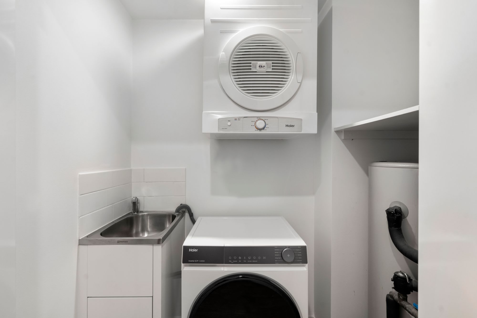 A compact laundry room with a front-loading washer and dryer, a stainless steel sink, and a ventilation unit above the dryer.