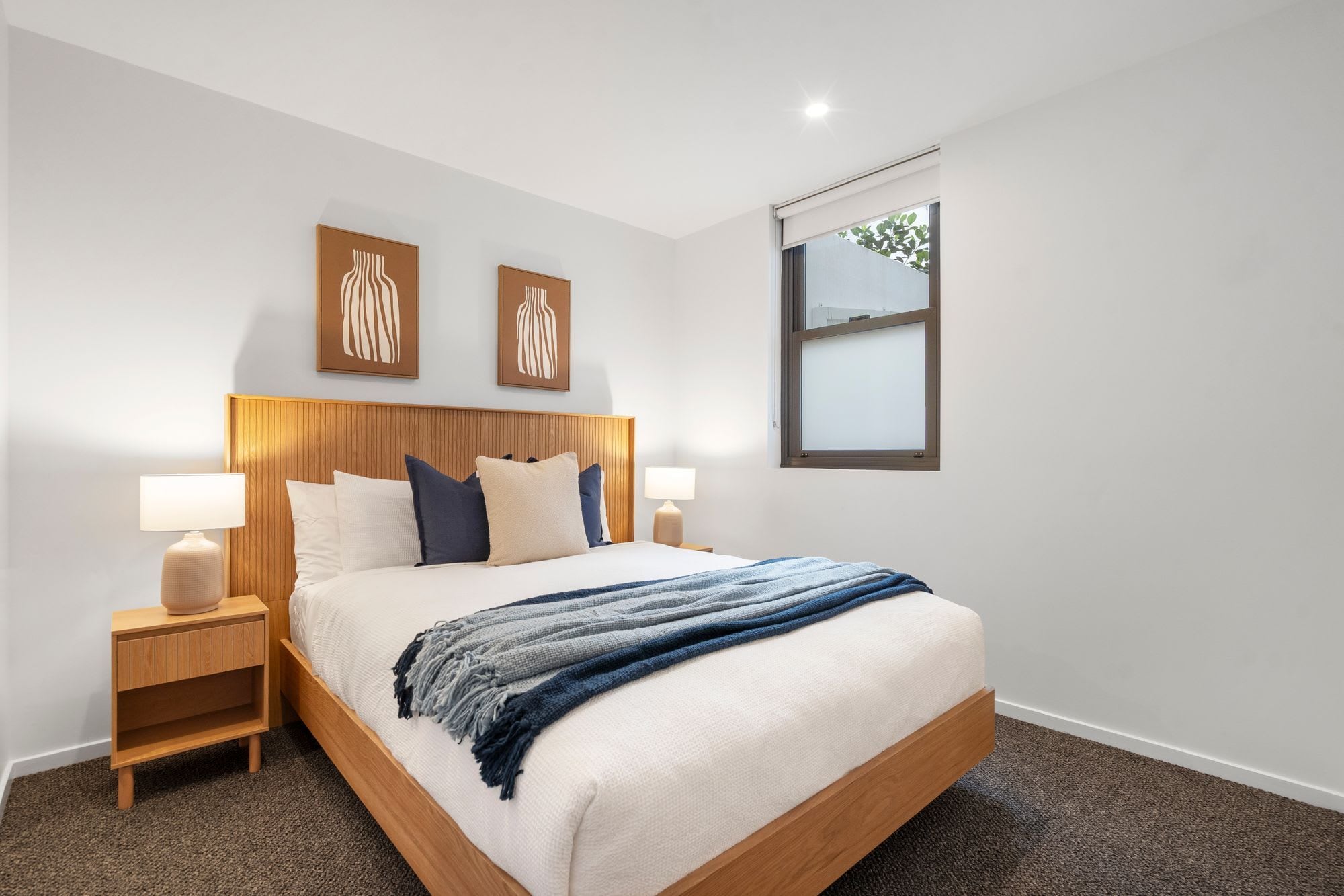 A hotel room with a wooden headboard, a bed with white bedding and blue accents, a nightstand with a lamp, and a window with a view.