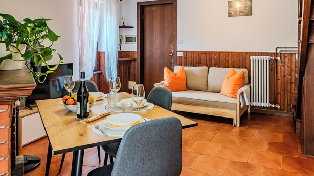 A cozy living room with a wooden floor, a sofa with orange cushions, a dining table set for four, and a potted plant. - Casa Gloria by Interhome (Porlezza)