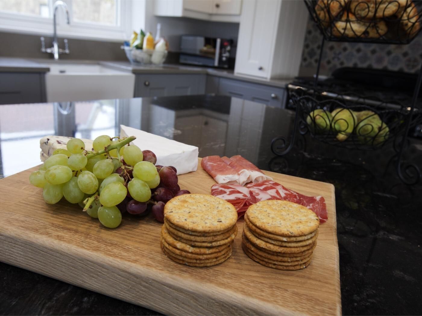 A wooden board with grapes, cheese, crackers, and sliced meat in a kitchen setting.
