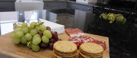 A wooden board with grapes, cheese, crackers, and sliced meat in a kitchen setting.