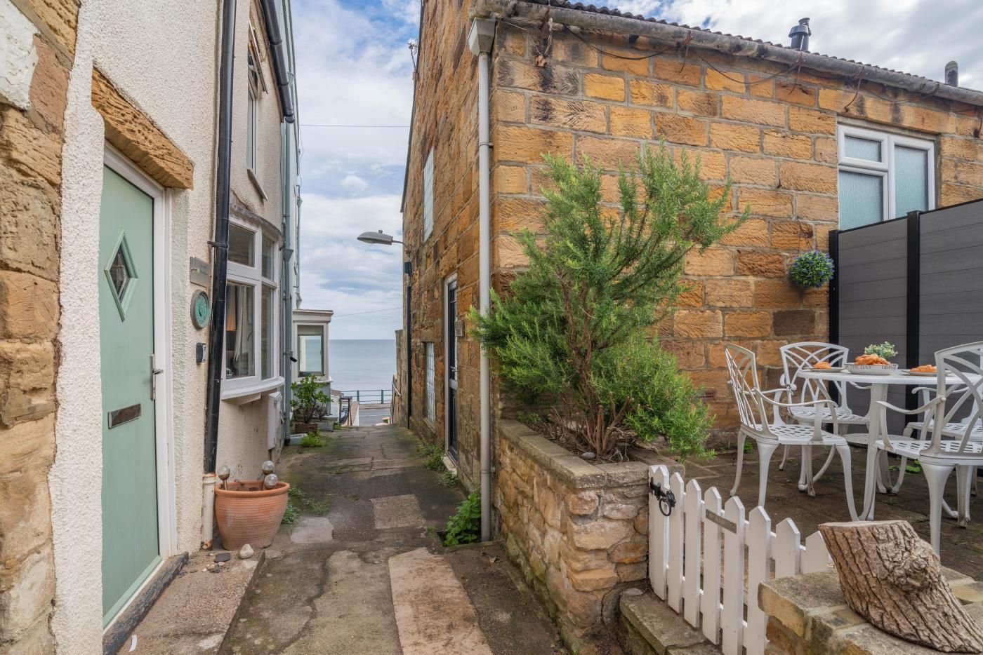 A narrow alleyway with stone buildings, a white picket fence, and a small outdoor seating area with chairs and a table.