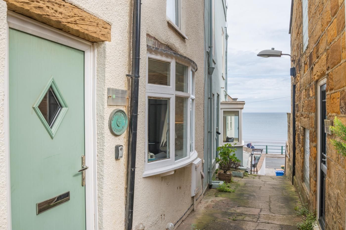 A narrow alleyway with a light green door, a window with a view of the sea, and a stone building on the right.