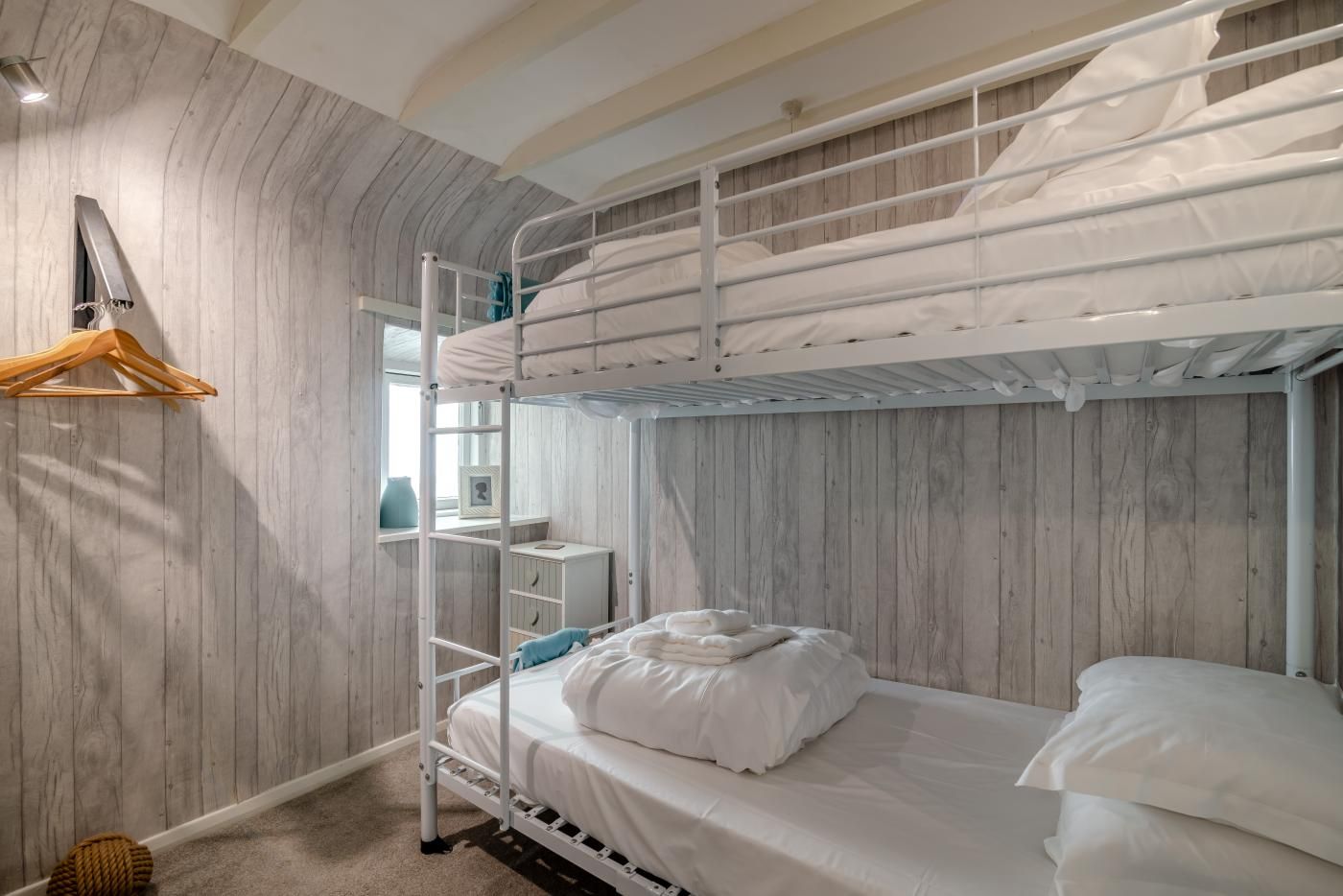 Habitación con literas, pared de madera, cama blanca, estante y perchero.