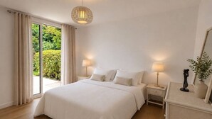 Une chambre à coucher avec un grand lit, deux tables de chevet, une lampe, un miroir et une vue sur la verdure à l’extérieur.