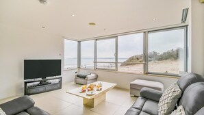 Ein modernes Wohnzimmer mit großem Fenster, einem Flachbildfernseher, einem Couchtisch mit Frühstücksangebot und einem grauen Sofa.
