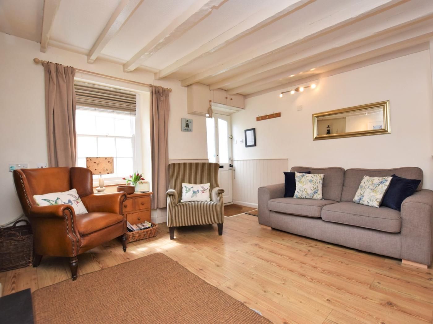 A living room with a brown leather armchair, a striped armchair, and a grey sofa. There is a wooden floor, a window with curtains, and a mirror on the wall.