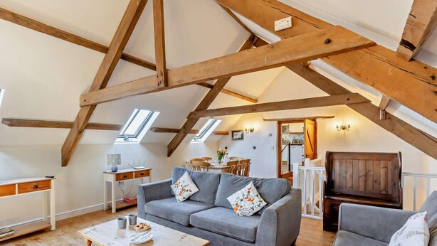Une pièce de style « loft », avec des poutres en bois apparentes, un canapé gris, une table basse en bois et un coin repas comprenant une table et des chaises blanches.