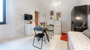 Habitació petita amb llit, escriptori amb cadira, televisió, radiador i taula baixa amb un ram de flors.