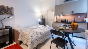 Habitació senzilla amb llit, escriptori amb làmpada, taula petita amb un bol de fruita i petita cuina.