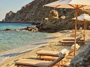 A beach with sun loungers and umbrellas, rocky cliffs in the background, and clear blue water.