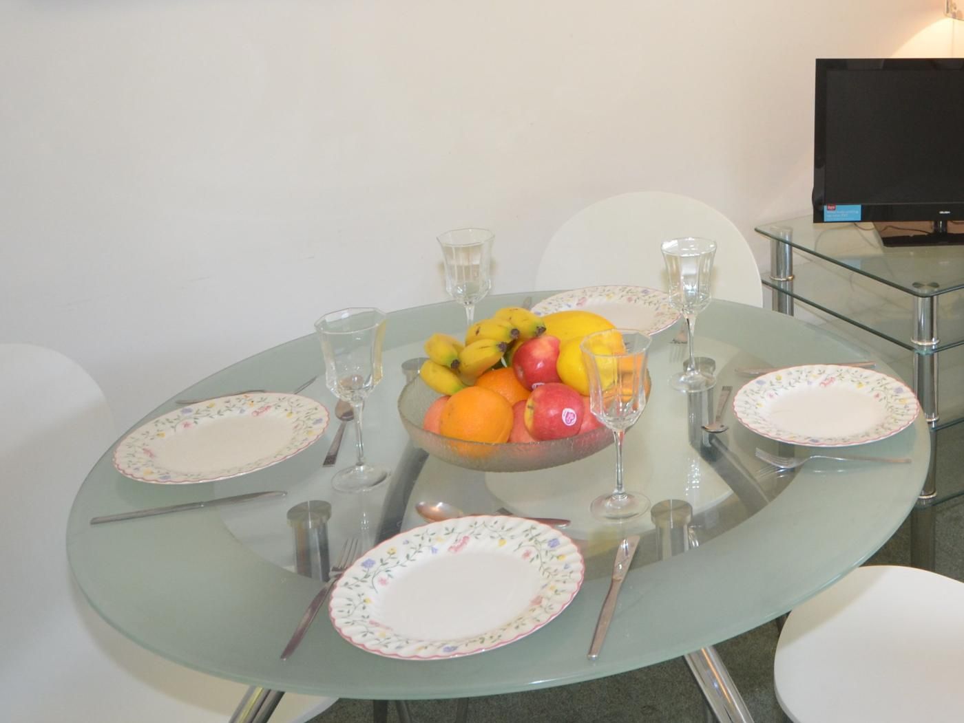 Une table ronde en verre, sur laquelle se trouvent un saladier de fruits, des assiettes, des couverts et des verres.