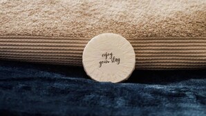 Ein Bett mit einer beige farbenen Decke und einem dunkelblauen Kopfkissen, ein runder Aufhänger mit dem Text 'enjoy your stay' auf einer strukturierten Oberfläche.