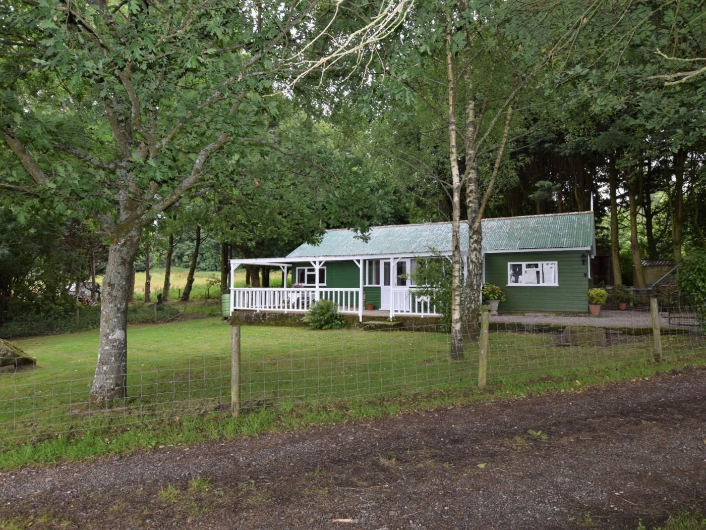 A green cottage with a porch, surrounded by trees and a fenced yard.