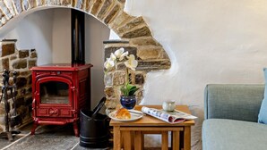 A cozy room with a red wood-burning stove, a stone fireplace, a wooden table with a plate, a cup, and a magazine. - 3 Bed in Bude (oc-heyle) (Launcells)