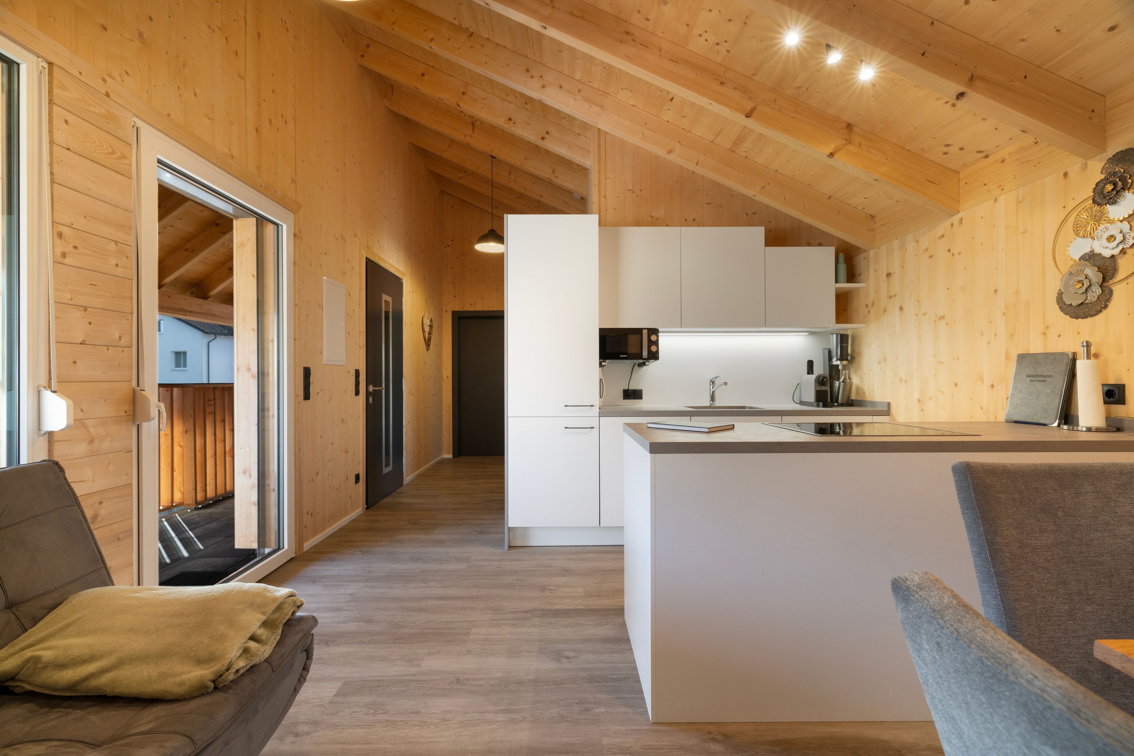 Eine moderne Küche mit einer zentralen Kochinsel, Holzbalken an der Decke und Blick in einen Wohnbereich mit einer Couch und einem Fenster.