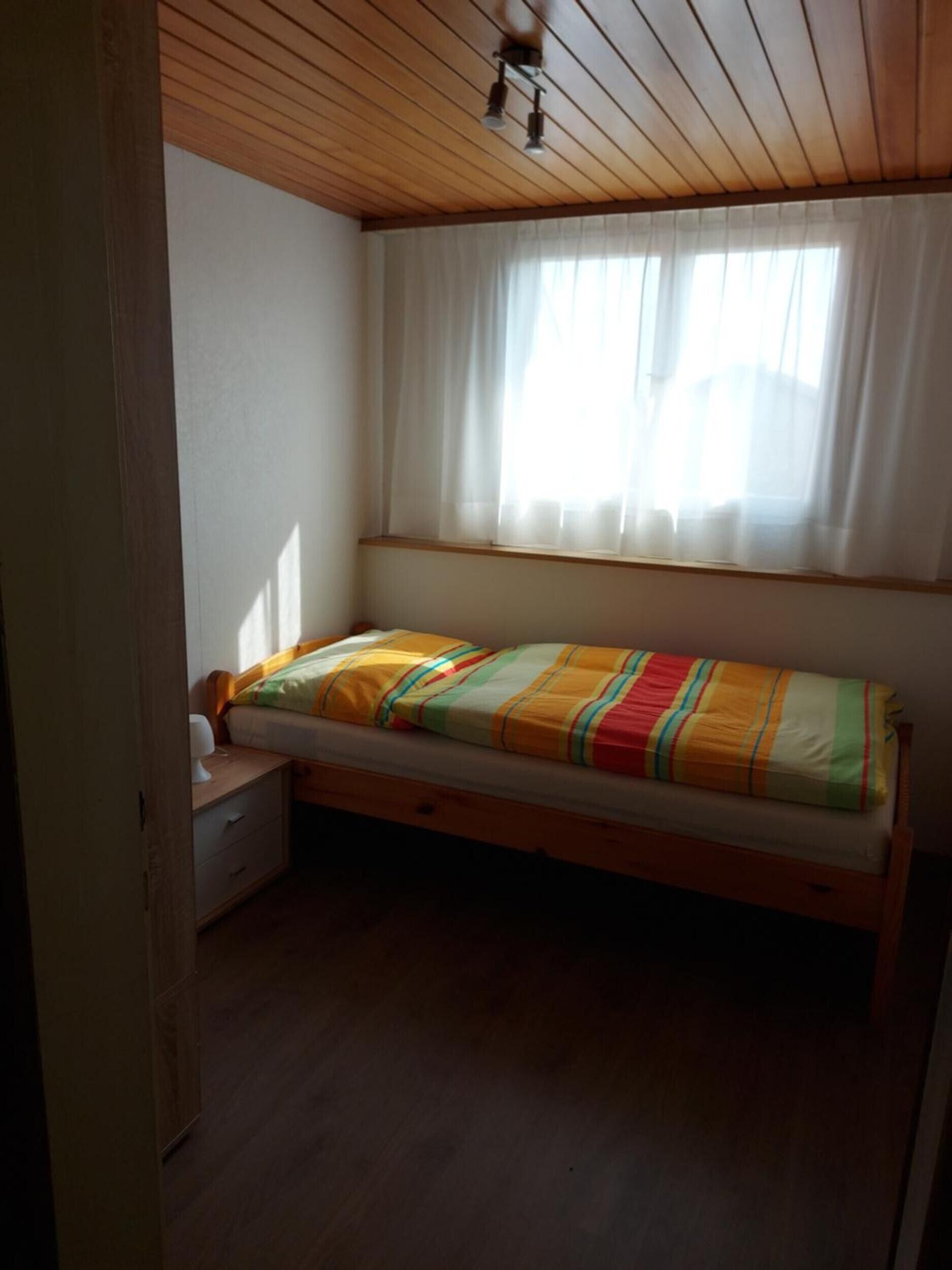 A small, single-bed room with a wooden ceiling, a window with sheer curtains, and a bedside table with a lamp.