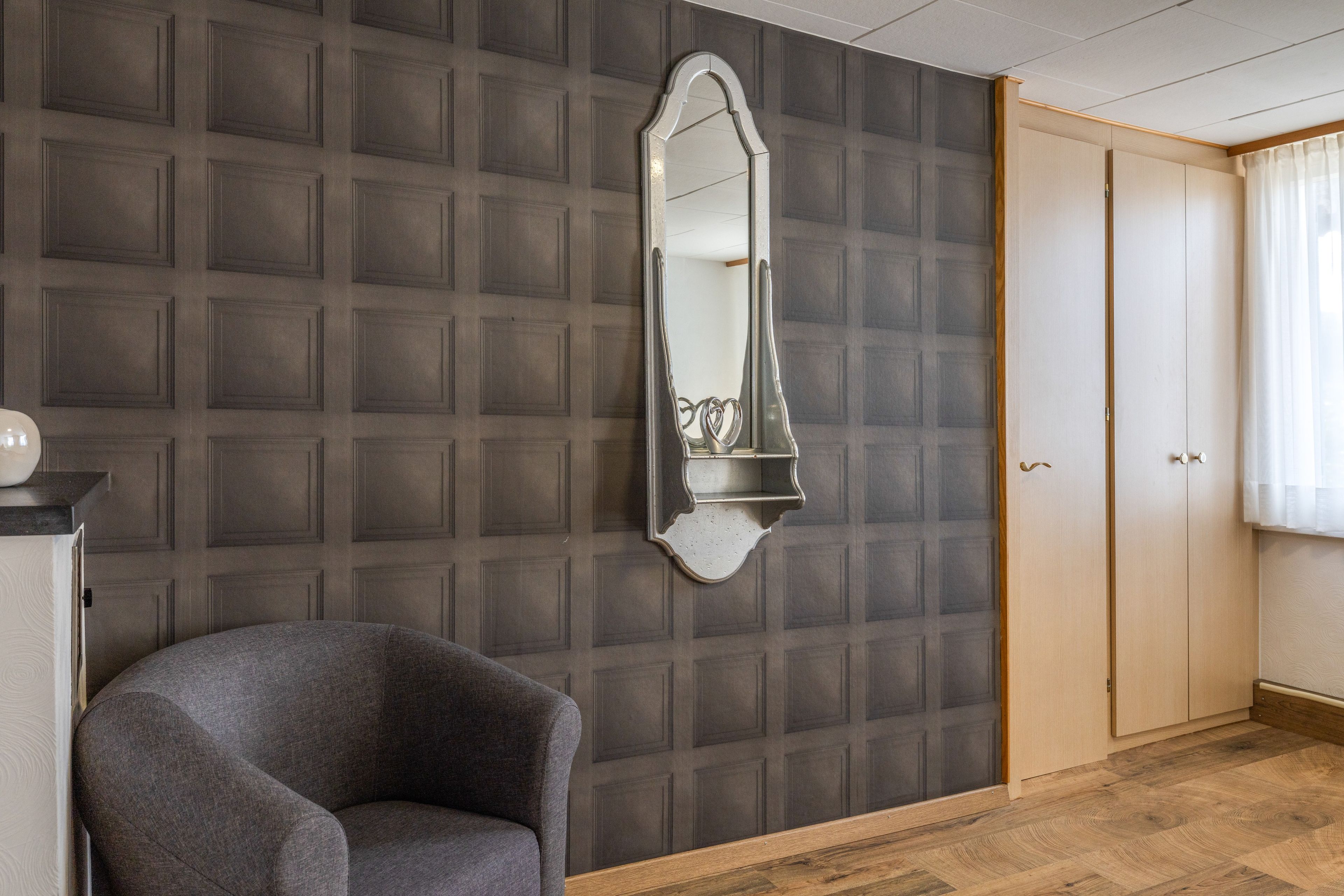 Een kamer met een behang, een spiegel, een stoel en een kledingkast.