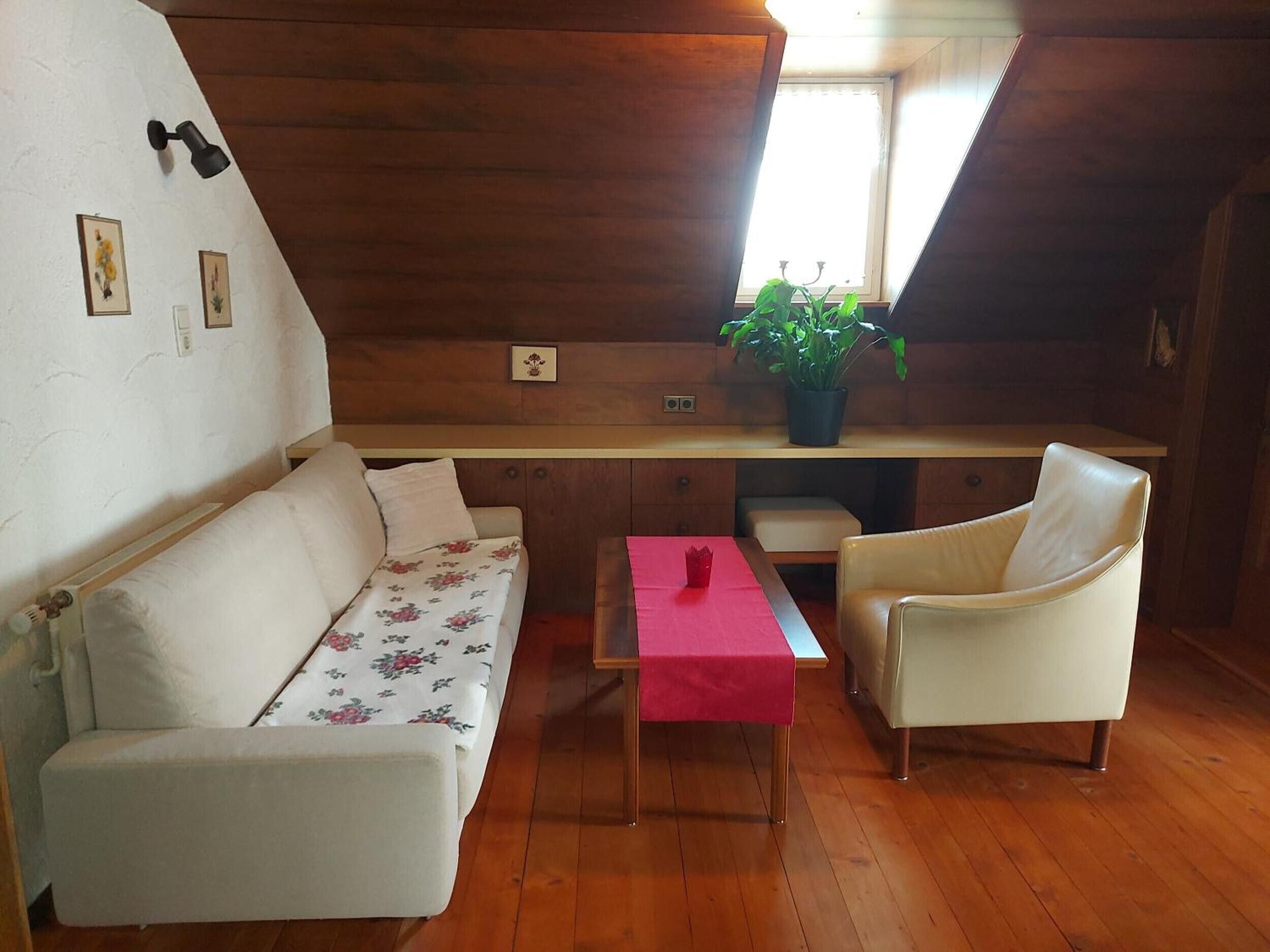 Ein gemütliches Zimmer mit Holzbalkendecke, einer mit Blumenmuster bezogenen Couch, einem Sessel, einem kleinen Tisch mit roter Tischdecke und einer Topfpflanze.