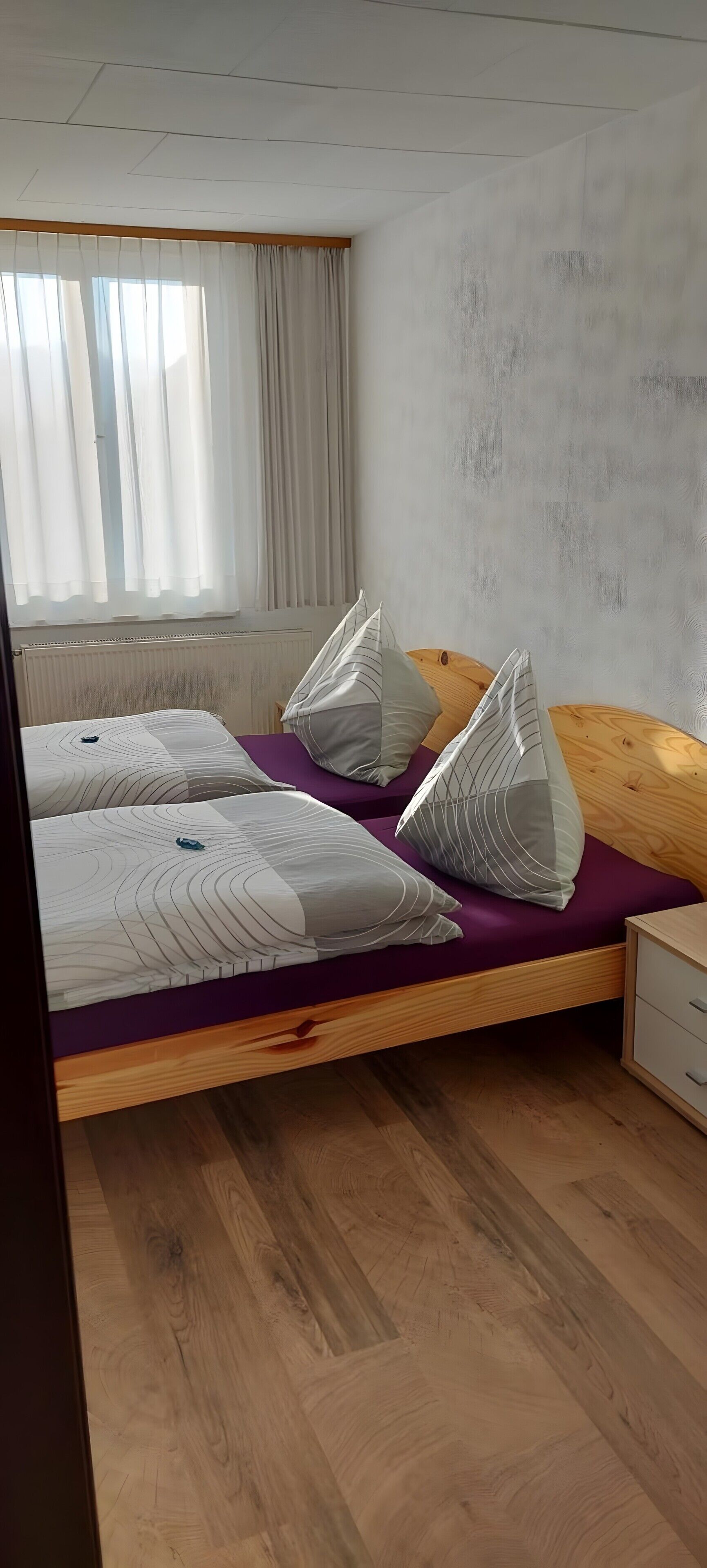 A bedroom with a bed, wooden headboard, a nightstand, and a window with curtains.