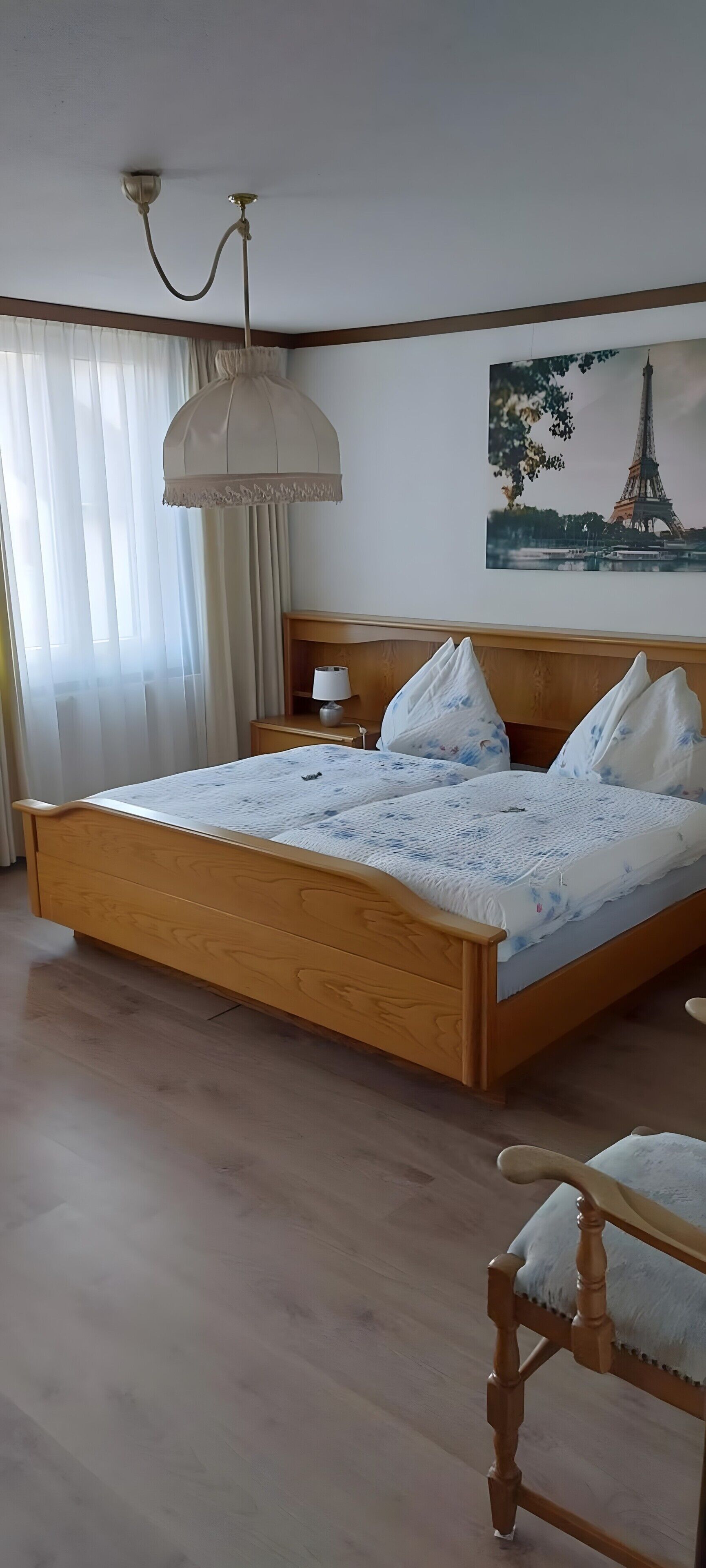 A bedroom with a wooden bed, a nightstand, a lamp, a chair, a ceiling light fixture, a window with curtains, and a picture of the Eiffel Tower on the wall.