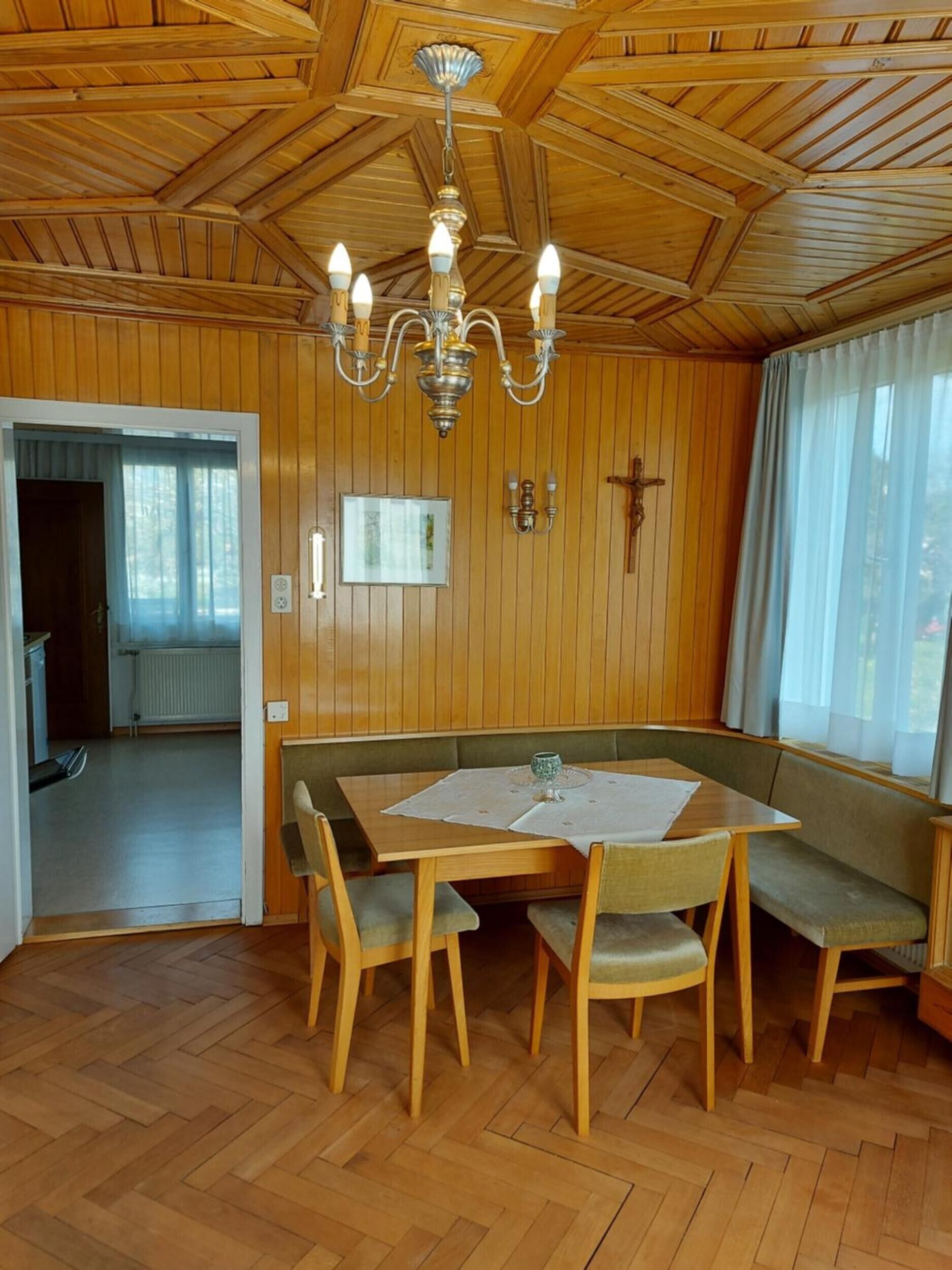 A wooden-paneled room with a dining table, benches, a chandelier, and a crucifix on the wall.
