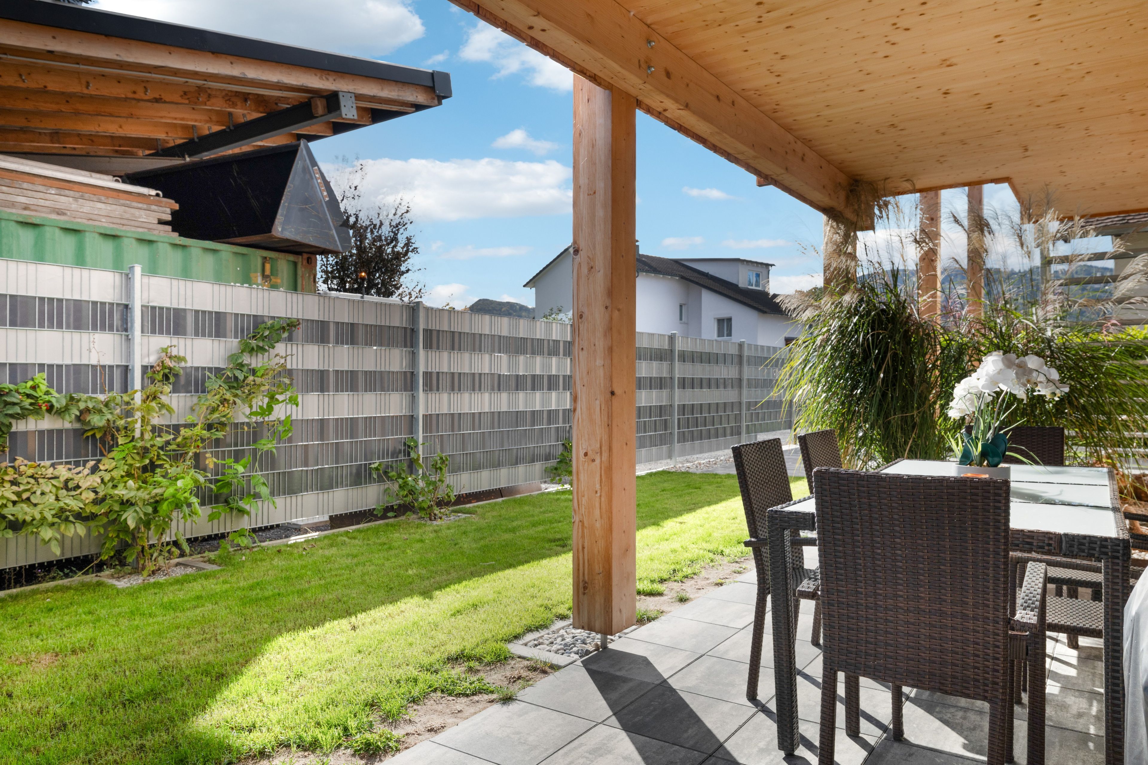 En terrasse med et bord og stole, en træpergola, et metalhegn og en græsplæne.