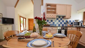 Un comedor con una mesa redonda dispuesta para cuatro personas, rodeada de sillas de madera. Se observa una cocina en el fondo con armarios de tonos claros y un salpicadero a cuadros azules.