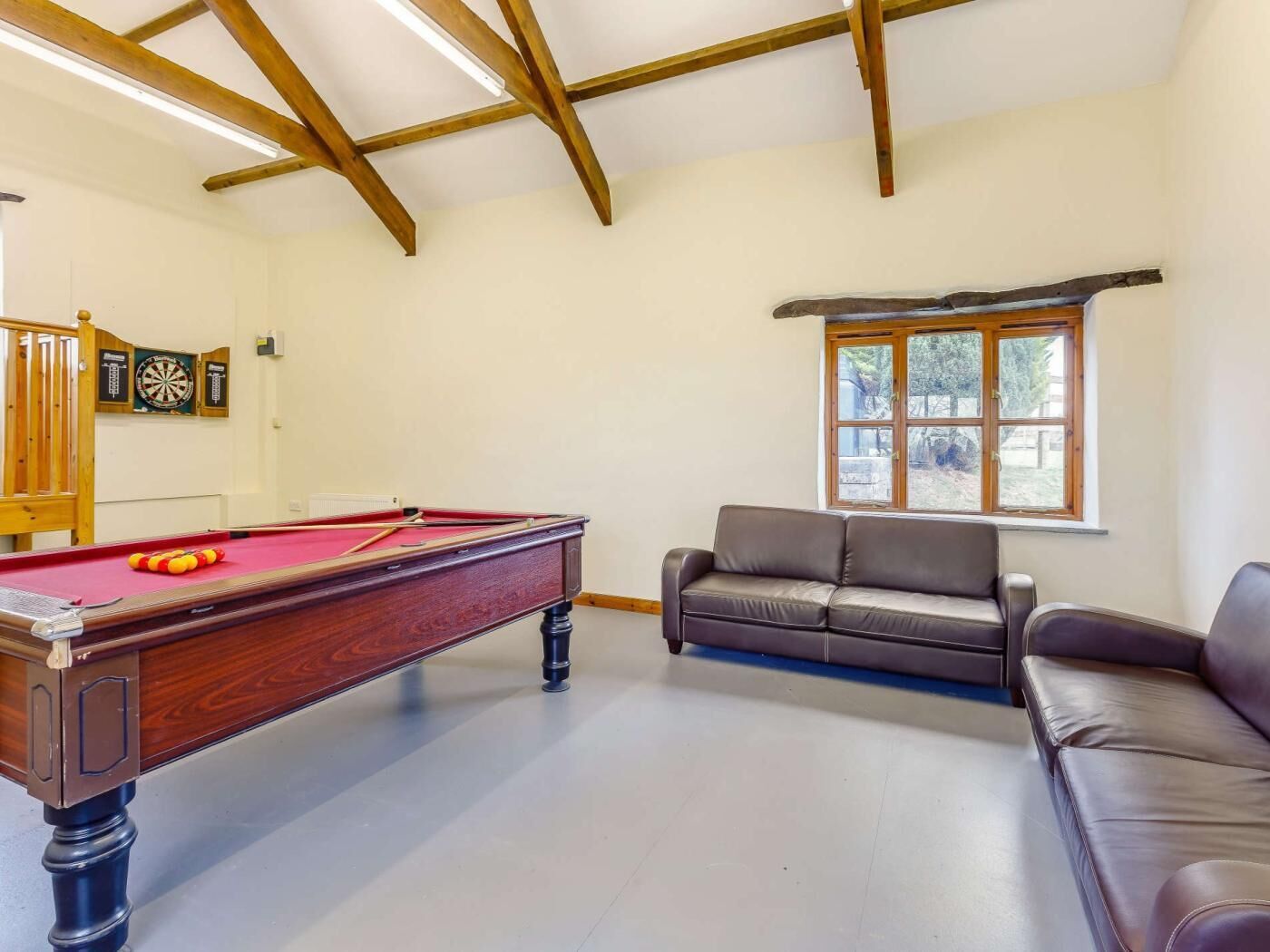 Une table de billard recouverte d’un feutre rouge, un canapé en cuir marron et une fenêtre donnant sur des arbres.