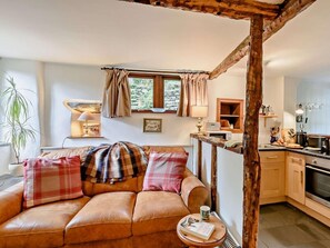A cozy living room with a leather sofa, a small table with a book and a cup, a kitchen area with a stove and cabinets, and a window with curtains. - 1 Bed in Nr Kendal (oc-lcc70) (Crosthwaite)