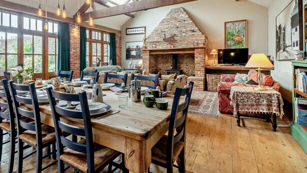 A dining area with a wooden table and chairs, a brick fireplace, and a television on a stand. - 5 Bed in Wiveton (oc-kt021) (Wiveton)