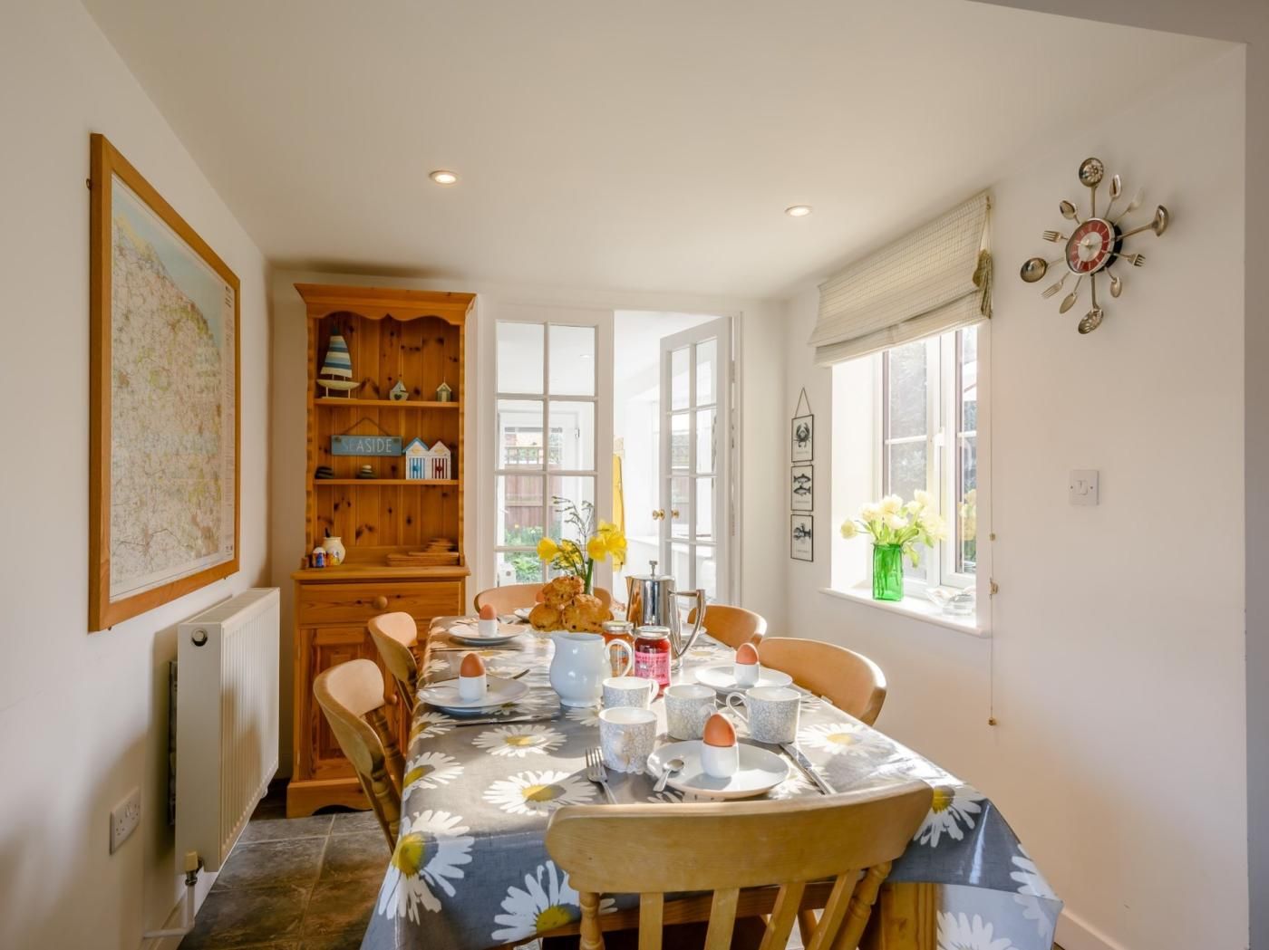 A dining area with a table set for four, wooden chairs, a bookshelf, and a window with a view.