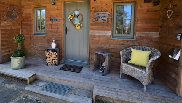 A wooden cabin with a front door, two windows, a wicker chair with a cushion, a small table with a vase, and a welcome sign. - 1 Bed in Thornton Watlass (oc-g0089) (Thornton Watlass)