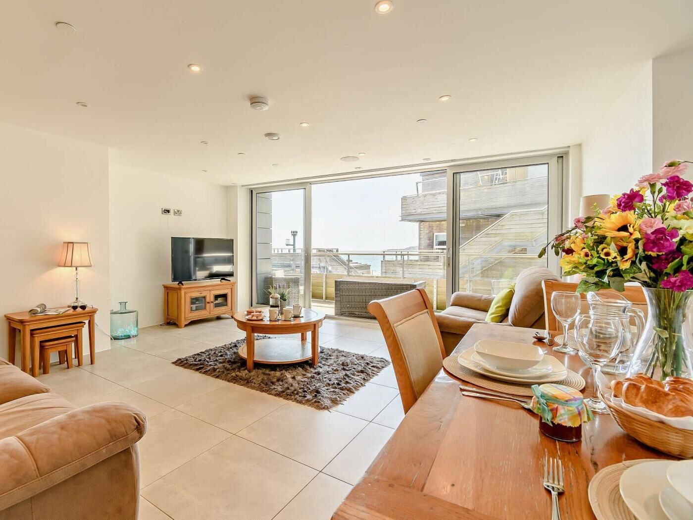 A modern living room with a TV, sofa, dining table, and a view of a building through large windows.