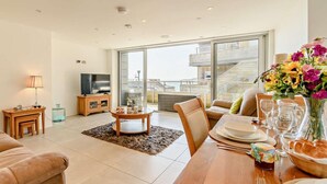 A modern living room with a TV, sofa, dining table, and a view of a building through large windows. - 2 Bed in Tenby (oc-fb290) (Tenby)