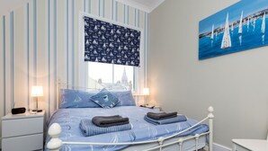 Een bed met een wit metalen frame, een raam met een geblindeerd rolgordijn, en een schilderij van zeilboten aan de muur.