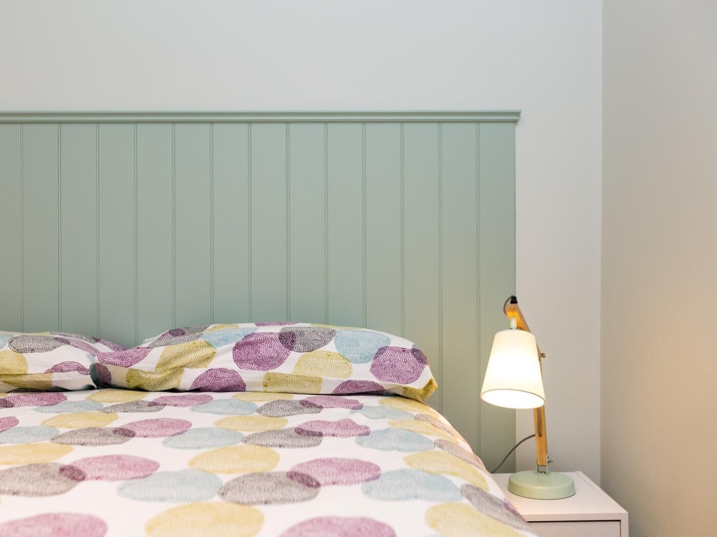 A bed with a colorful circular pattern bedspread and pillows, a bedside lamp, and a green-painted headboard.