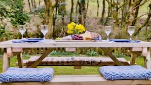 A picnic setup with a wooden table, two wine glasses, a cheese platter, and a vase of yellow flowers. - 3 Bed in Cleator (oc-sz392) (Ennerdale Bridge)