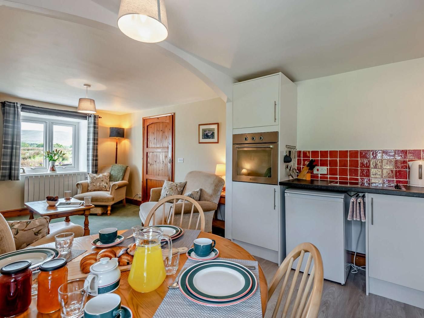 A cozy living room with a dining table set for breakfast, a kitchen with modern appliances, and a view through a window.