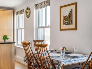 A dining area with a table set for two, two wooden chairs, a clock, and a framed picture on the wall. - 2 Bed in Tenby (oc-fb115) (Tenby)