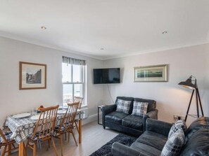 A living room with a dining area, a black leather sofa, a flat-screen TV mounted on the wall, and a framed picture above the sofa. - 2 Bed in Tenby (oc-fb115) (Tenby)