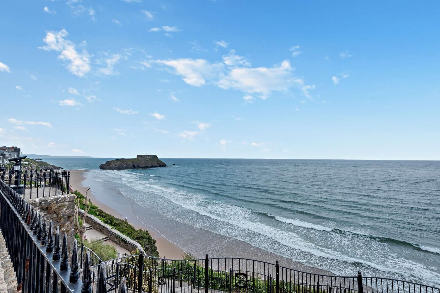 A coastal view with a stone staircase leading to a beach, a fortified structure on a small island, and a clear sky.