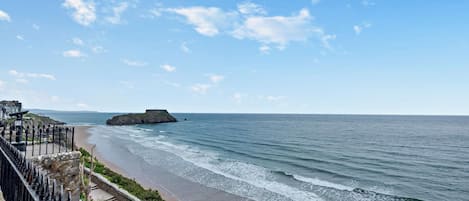 A coastal view with a stone staircase leading to a beach, a fortified structure on a small island, and a clear sky.