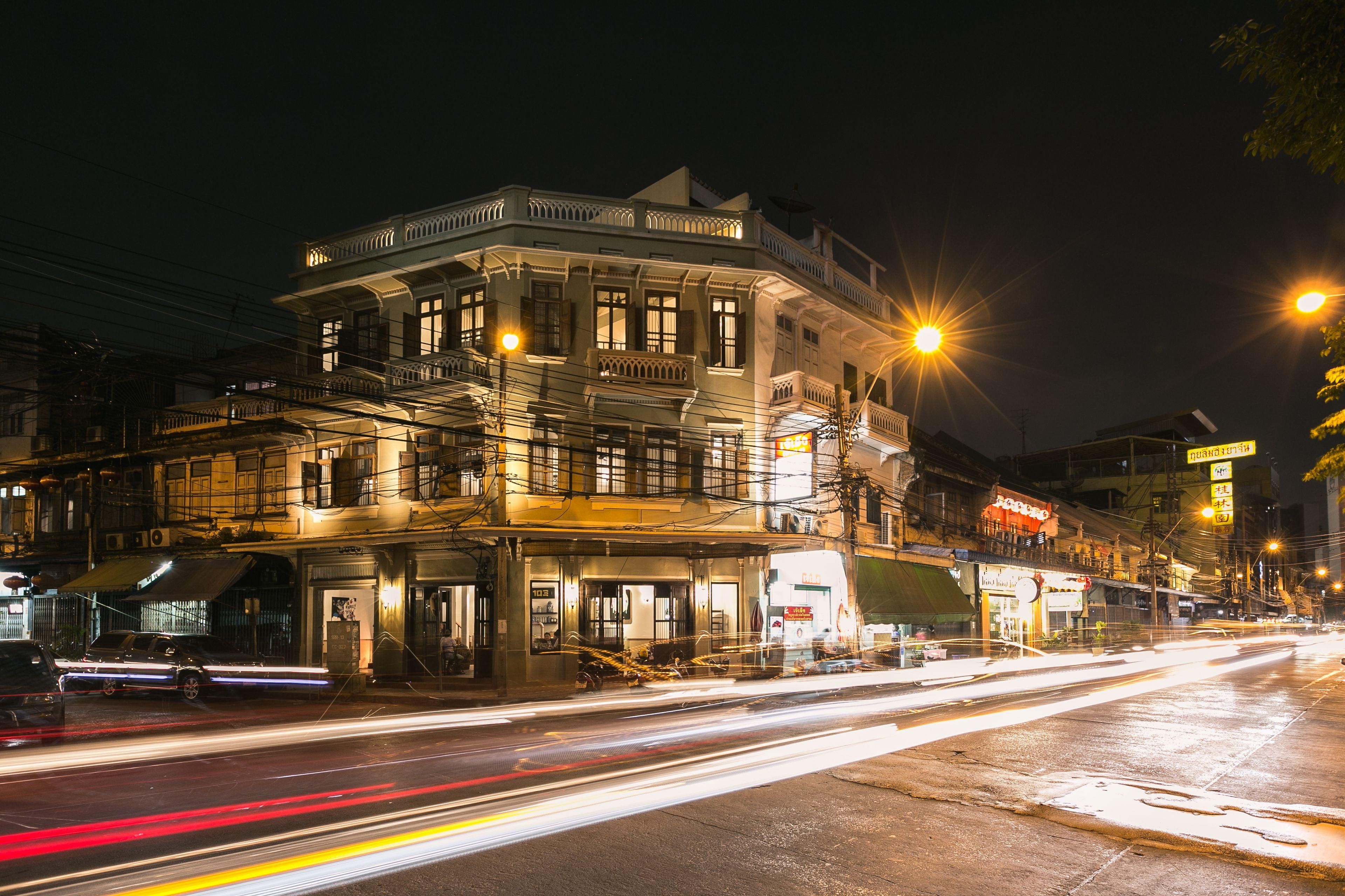 A multi-story building with a balcony on the second floor, a staircase on the third floor, and a street in front with light trails from moving vehicles.