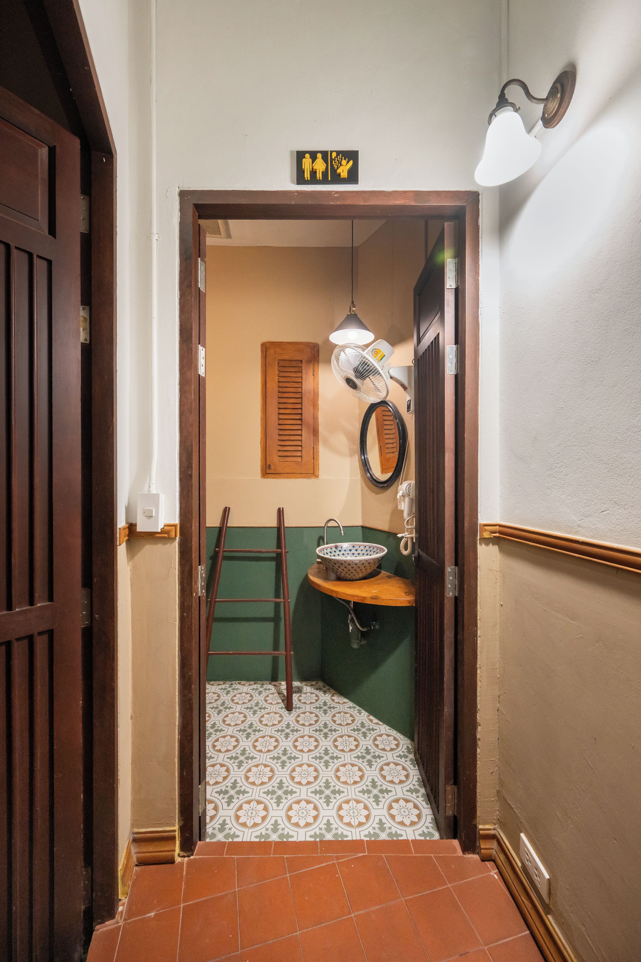 A narrow hallway with a wooden door, a white wall with a black and yellow sign, a white wall-mounted fan, a wooden ladder, and a green tiled floor.