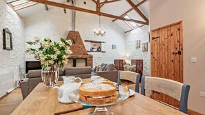 Ein rustikales Interieur mit freiliegenden Holzbalken, einem Kamin aus Backstein, einem Esstisch mit Kuchen und einer gemütlichen Sitzecke.